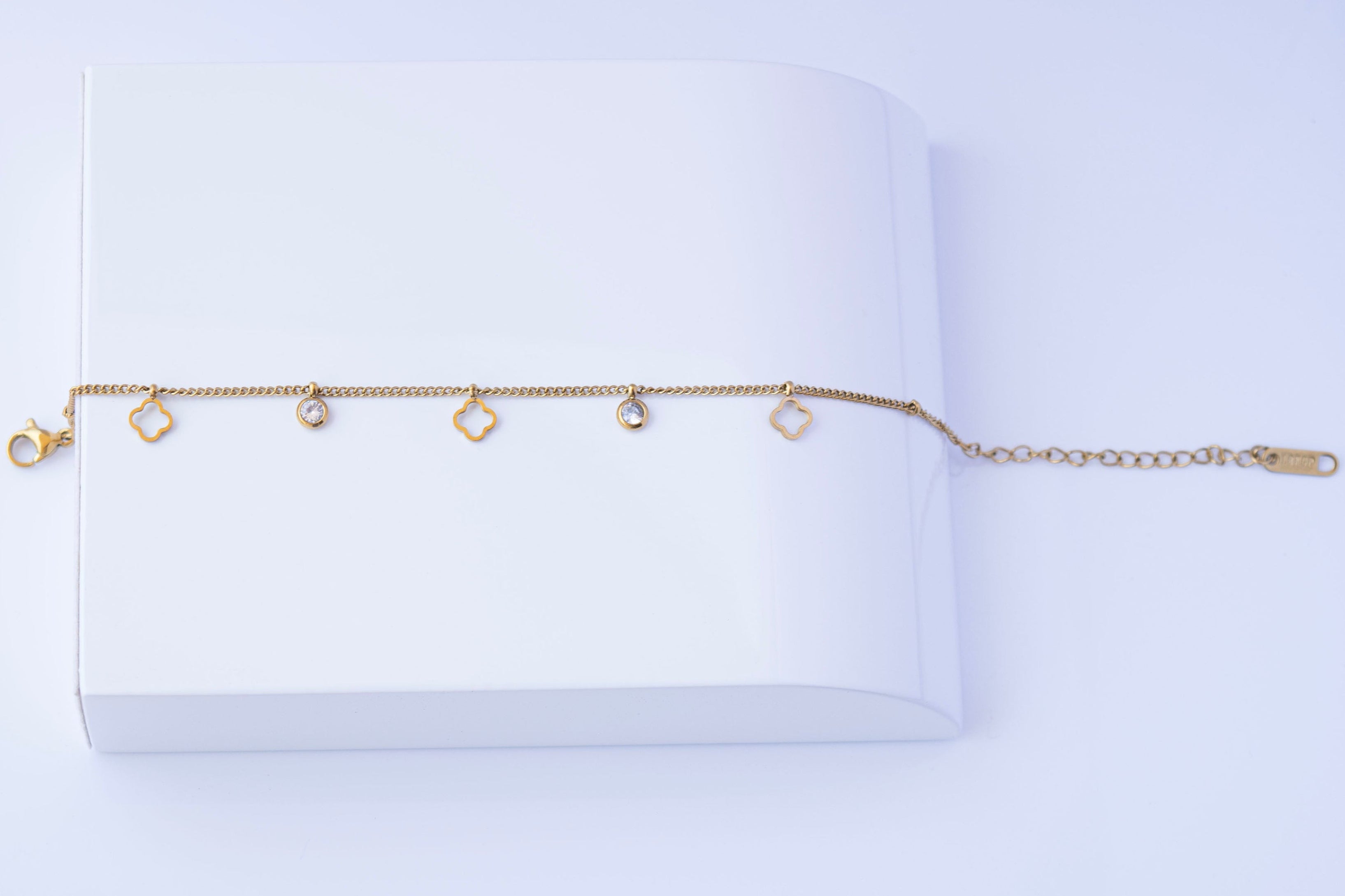 A delicate stainless steel chain bracelet with three small open diamond-shaped charms and two small round bezel-set clear stones horizontally on a white book-like prop against a light background.